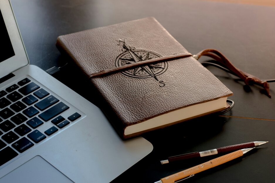 Laptop, zwei Stifte und in braunem Leder eingebundenes Buch mit Kompass auf Tisch | © Unsplash