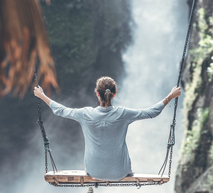 Urlauberin sitzt auf Holzschaukel vor Wasserfall | © Pexels