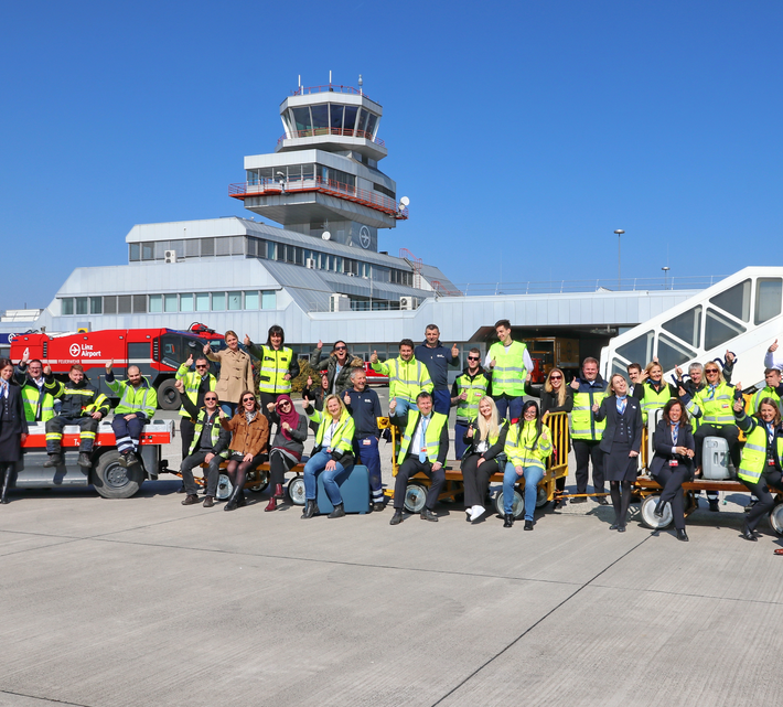 Flughafenmitarbeiter sitzen winkend auf Gepäckswagerl am Vorfeld vor dem Tower | © Linz Airport