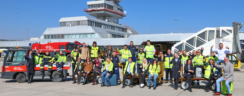 Flughafenmitarbeiter sitzen winkend auf Gepäckswagerl am Vorfeld vor dem Tower | © Linz Airport