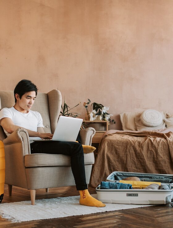 Junger Mann mit Laptop am Schoß sitzt zwischen Koffern und Globus in einem Ohrensessel | © Pexels