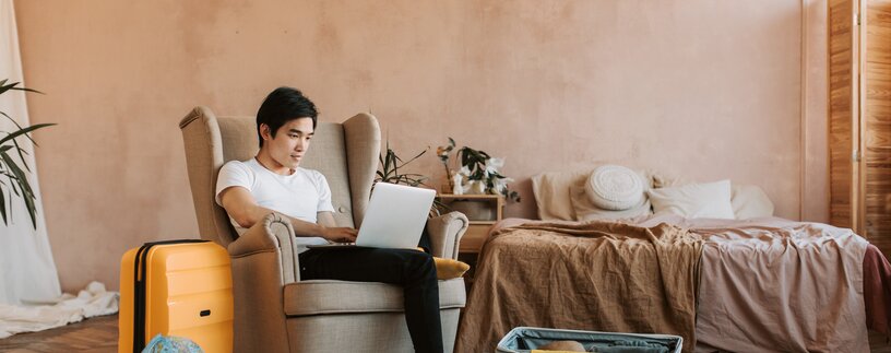 Junger Mann mit Laptop am Schoß sitzt zwischen Koffern und Globus in einem Ohrensessel | © Pexels