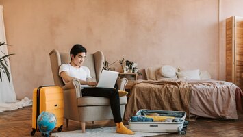 Junger Mann mit Laptop am Schoß sitzt zwischen Koffern und Globus in einem Ohrensessel | © Pexels