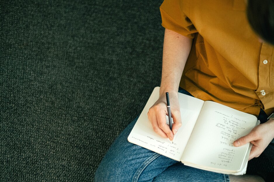 Junger Mann im Schneidersitz notiert eine Liste in ein Notizheft | © Pexels
