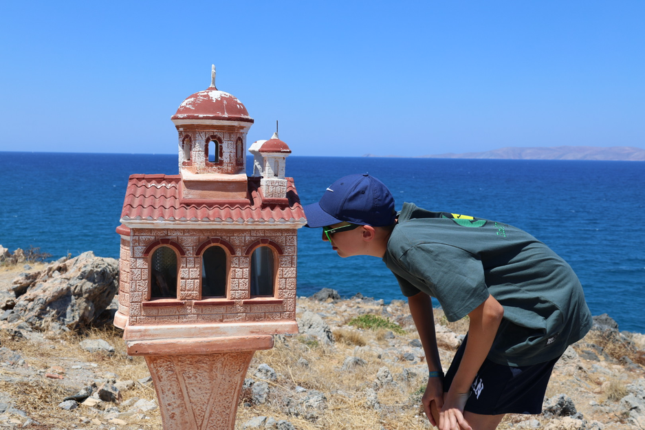 Junge sieht in das Innere einer kleinen griechischen Kirche auf einem Ständer vor Meer | © Linz Airport