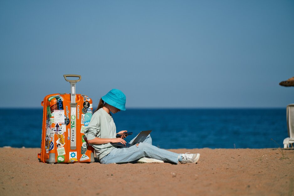 Mädchen mit Laptop und Handy sitzt angelehnt an einem orangen Koffer am Strand | © Unsplash
